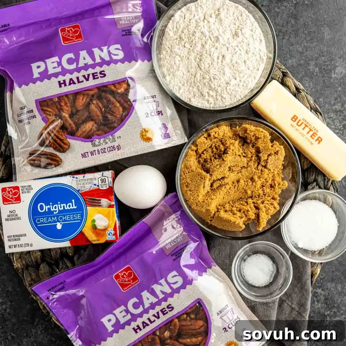Ingredients for baking include pecans, flour, butter, cream cheese, egg, brown sugar, and salt on a dark surface.