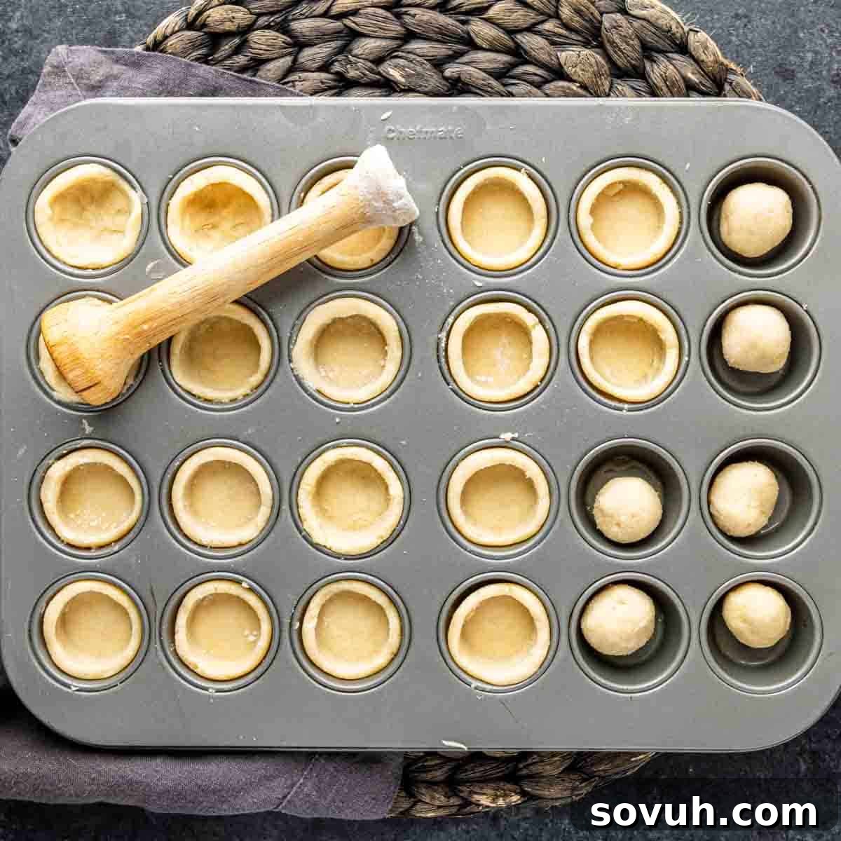 Mini tart dough in a muffin pan, with dough balls and a wooden tamper on top.