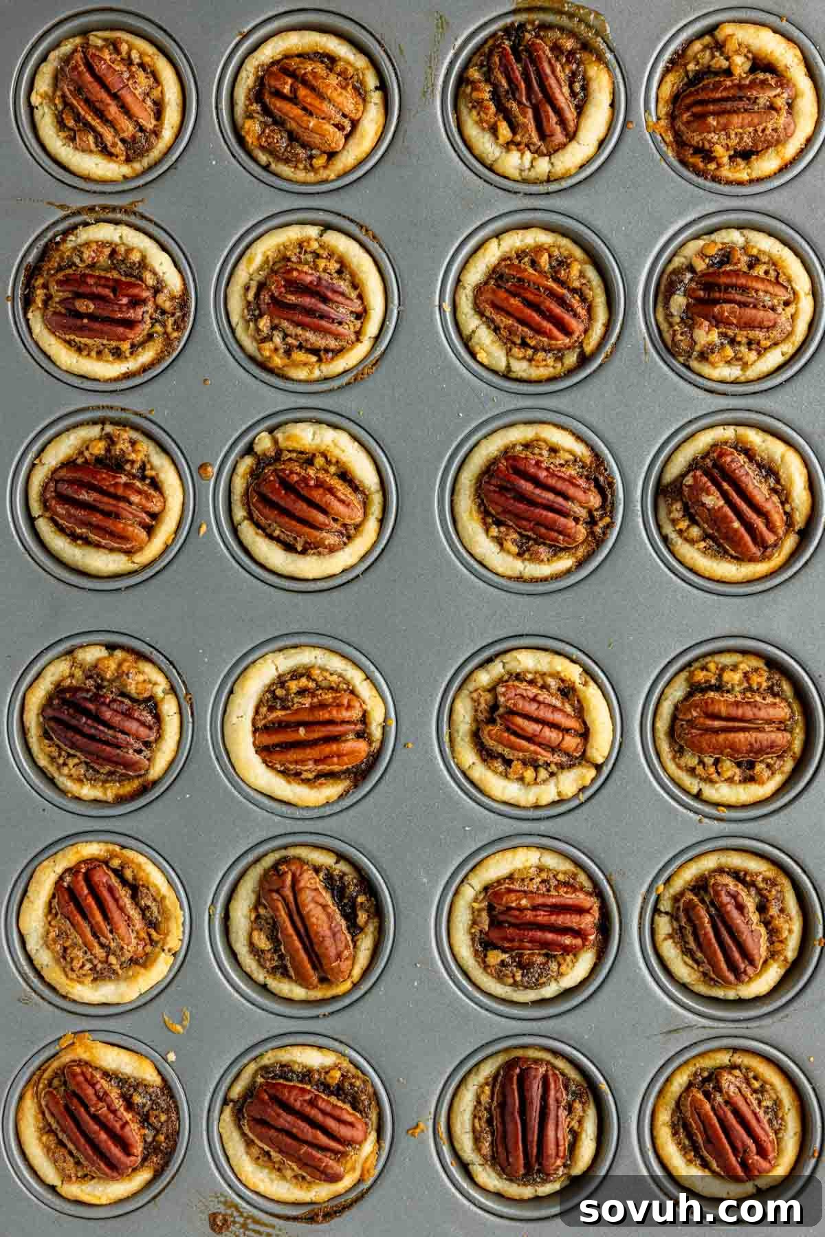 A baking tray with 24 mini pecan pies arranged in rows, each topped with a pecan half.