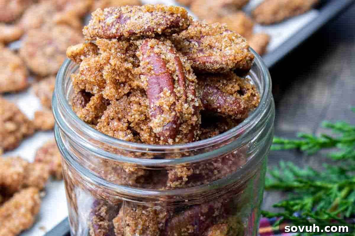 A jar filled with candied pecans sits on a table, with more pecans scattered on a tray in the background.