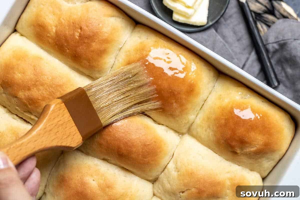 Freshly baked dinner rolls being brushed with melted butter in a baking dish, with a butter dish and knife visible in the background.