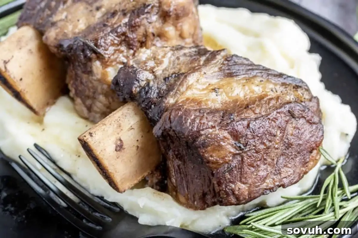 Braised beef short ribs served on a bed of mashed potatoes with a sprig of rosemary on the side, and a fork on a black plate.