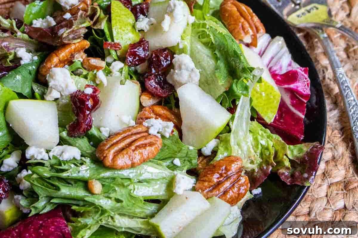 A fresh salad with mixed greens, pear slices, pecans, crumbled cheese, and dried cranberries in a black bowl on a woven mat.
