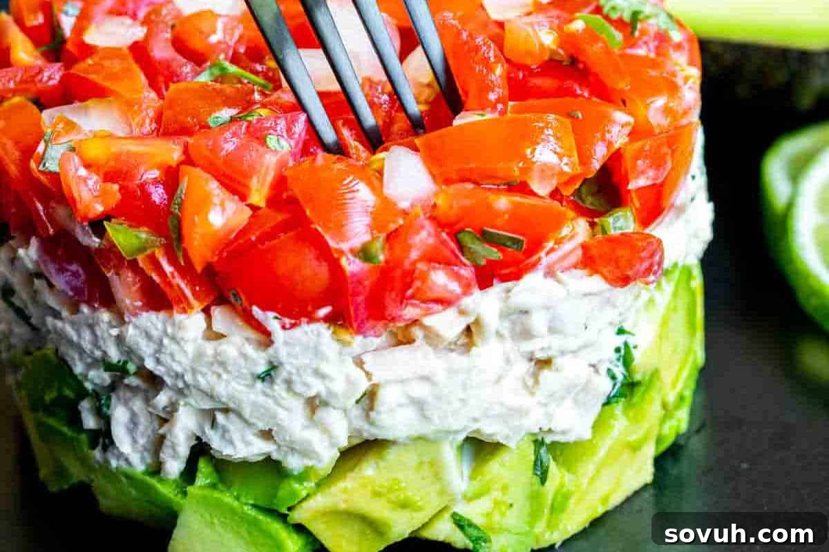 Layers of diced avocado, tuna salad, and chopped tomatoes with herbs. A fork is piercing the top layer. Lime slices are in the background.