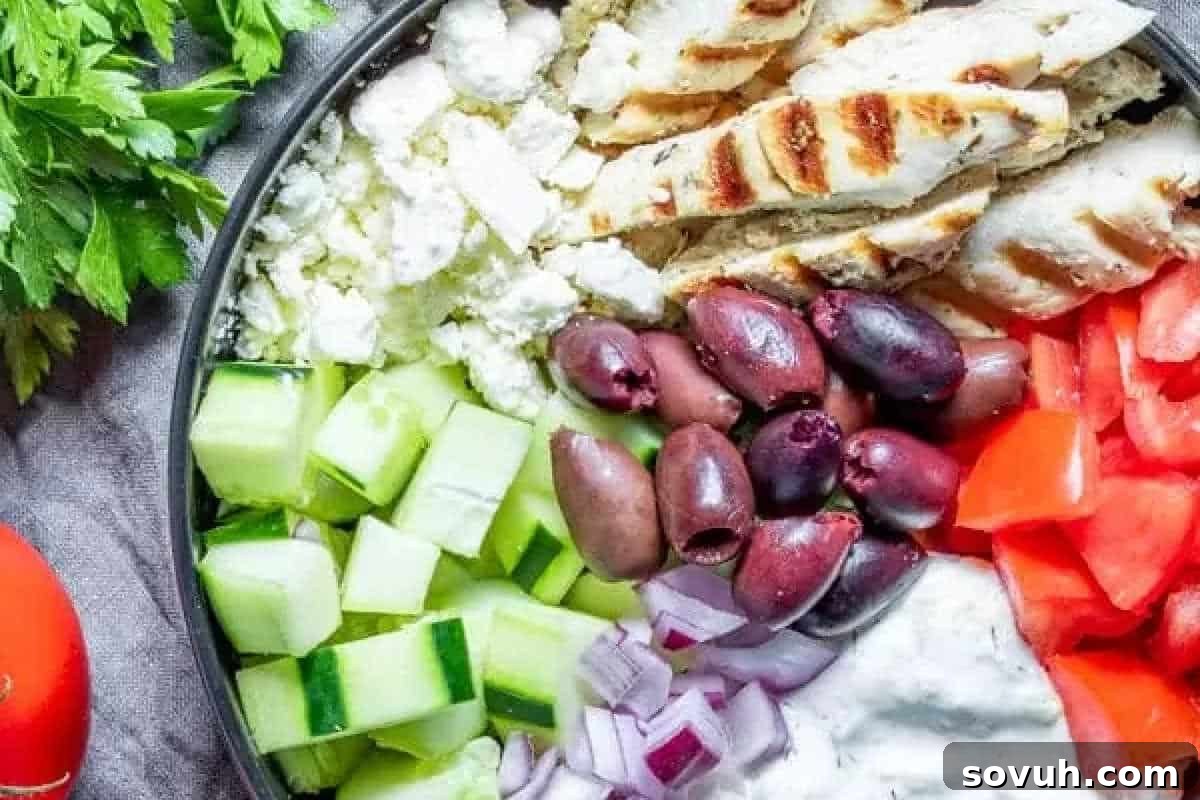 Bowl with grilled chicken, feta, cucumber, olives, diced tomato, onion, and tzatziki sauce. Fresh parsley and tomatoes are on the side.