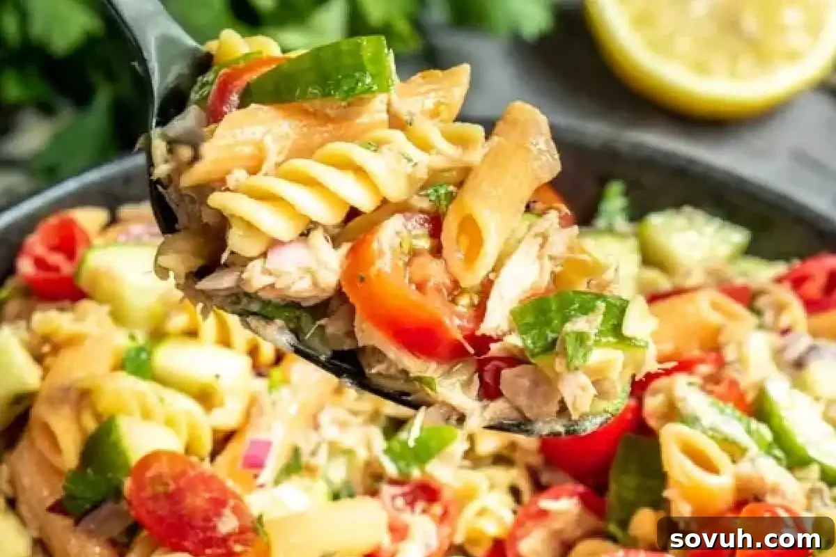 Close-up of a spoonful of colorful pasta salad with cherry tomatoes, green peppers, red onions, and herbs. A lemon slice is visible in the background.