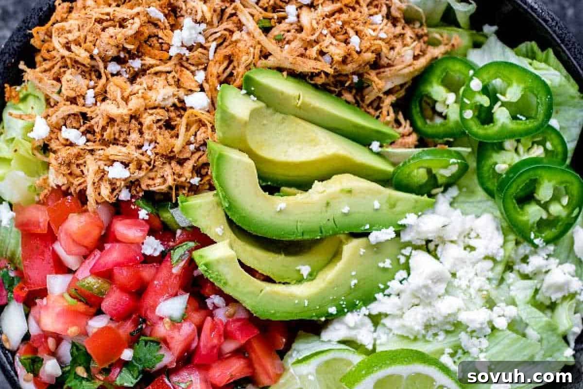A bowl with shredded chicken, sliced avocado, jalapeños, diced tomatoes, lettuce, and crumbled cheese.
