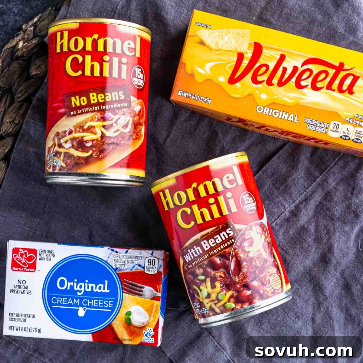Essential ingredients for chili cheese dip: cans of chili, a block of Velveeta cheese, and a package of cream cheese, neatly arranged on a dark surface.