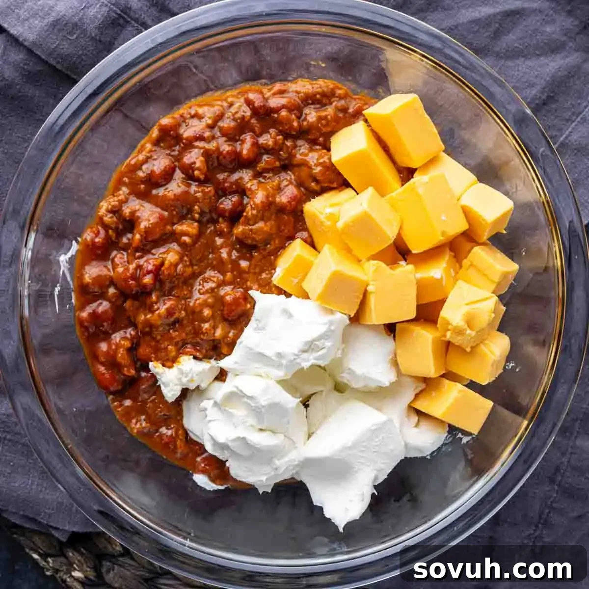 A microwave-safe bowl filled with chili, cubed Velveeta cheese, and softened cream cheese, ready to be melted.