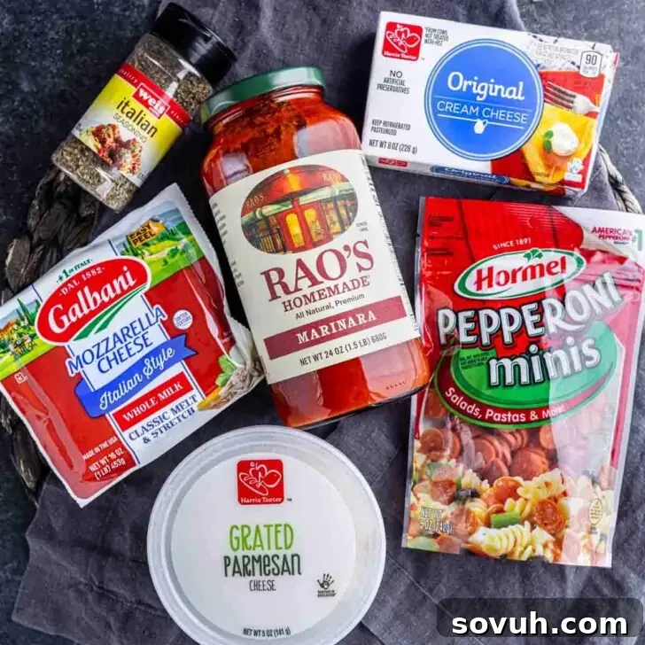 Ingredients for pizza dip: Italian seasoning, cream cheese, shredded mozzarella, marinara sauce, grated parmesan, and mini pepperoni arranged on a dark surface.