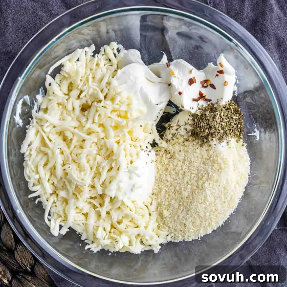 A glass bowl containing shredded cheese, sour cream, cream cheese, grated parmesan, dried herbs, and red pepper flakes on a dark surface, ready to be mixed.