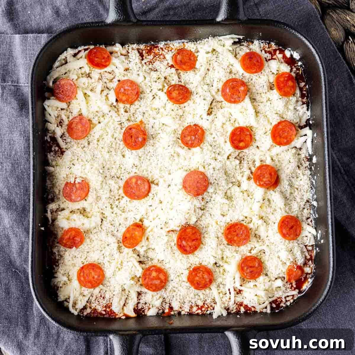 A square baking pan containing an uncooked pizza dip, topped with pepperoni slices and cheese, ready for the oven.