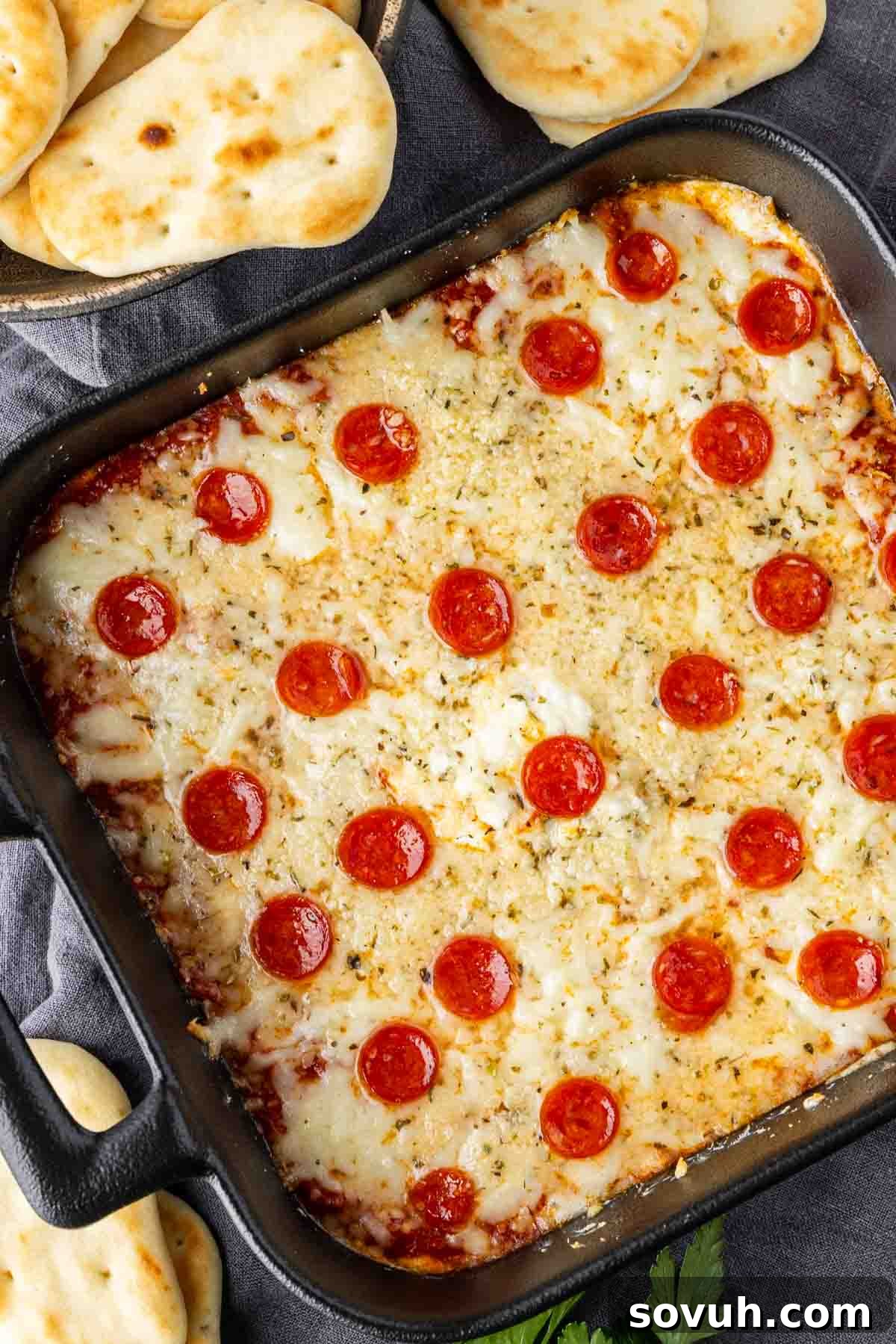 A rectangular pepperoni pizza dip in a black baking dish, topped with melted cheese and pepperoni, surrounded by pieces of toasted flatbread on a rustic surface.
