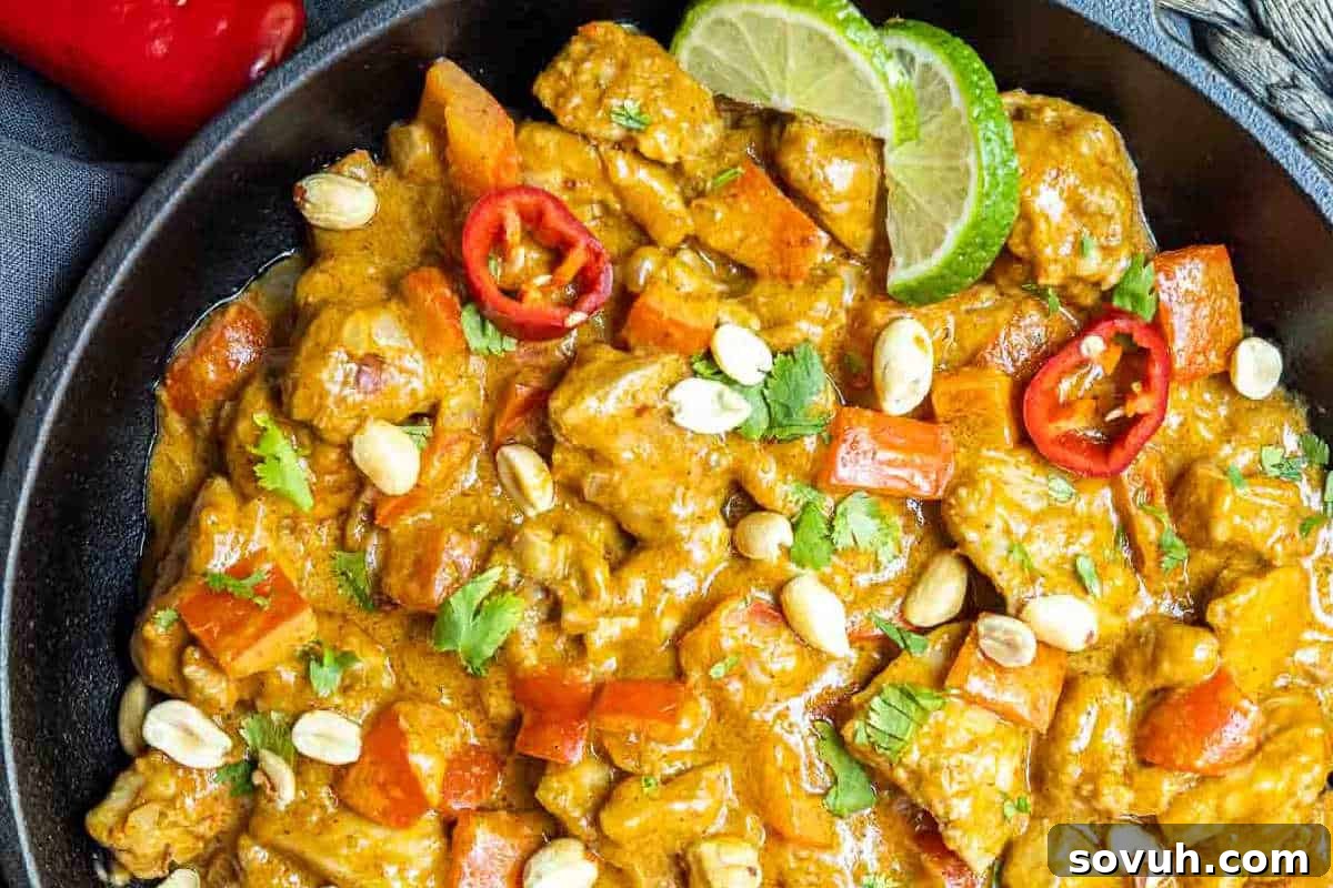 A vibrant bowl of Thai curry with tender chicken pieces, bright red peppers, crushed peanuts, a slice of fresh lime, and a sprinkle of cilantro garnish.