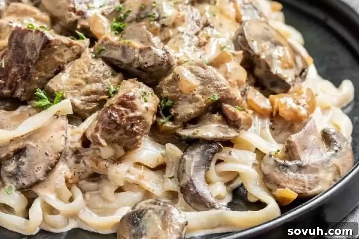 A savory plate of creamy pasta generously topped with perfectly sliced mushrooms and tender pieces of beef, beautifully garnished with a sprinkle of chopped fresh herbs.