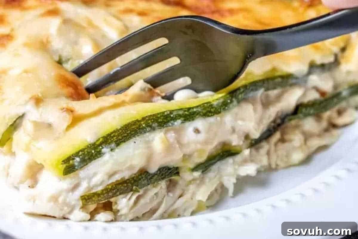 A fork elegantly cuts into a hearty slice of zucchini and chicken lasagna, perfectly presented on a pristine white plate, showcasing its layers.