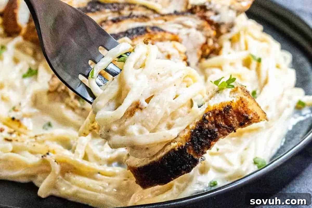 A fork gracefully lifts a generous portion of creamy fettuccine Alfredo, intertwined with a succulent piece of grilled chicken, from a pristine white plate.