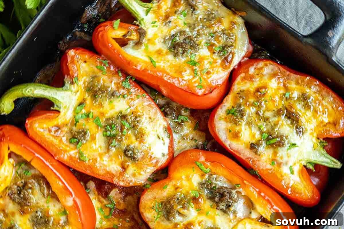 Vibrant red bell pepper halves are generously stuffed with a creamy cheese and meat mixture, then topped with bubbly melted cheese and garnished with fresh chopped herbs, presented in a sleek black baking dish.