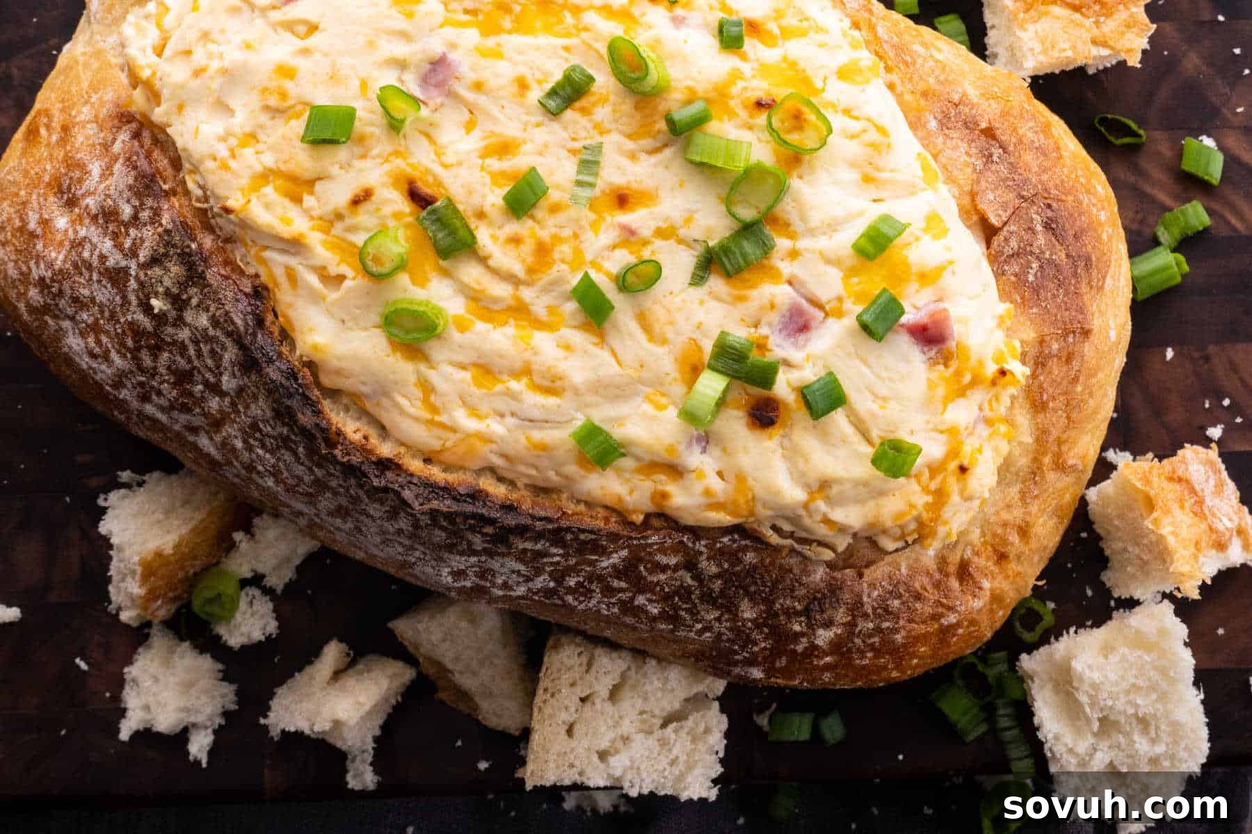 Loaf of bread hollowed and filled with a creamy dip, topped with chopped green onions, surrounded by bread pieces on a dark surface.