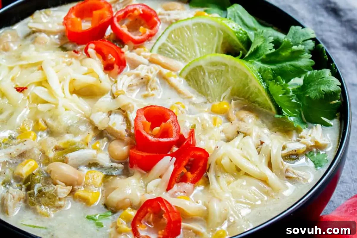 A bowl of creamy soup with shredded chicken, beans, corn, sliced red peppers, lime wedges, grated cheese, and cilantro.