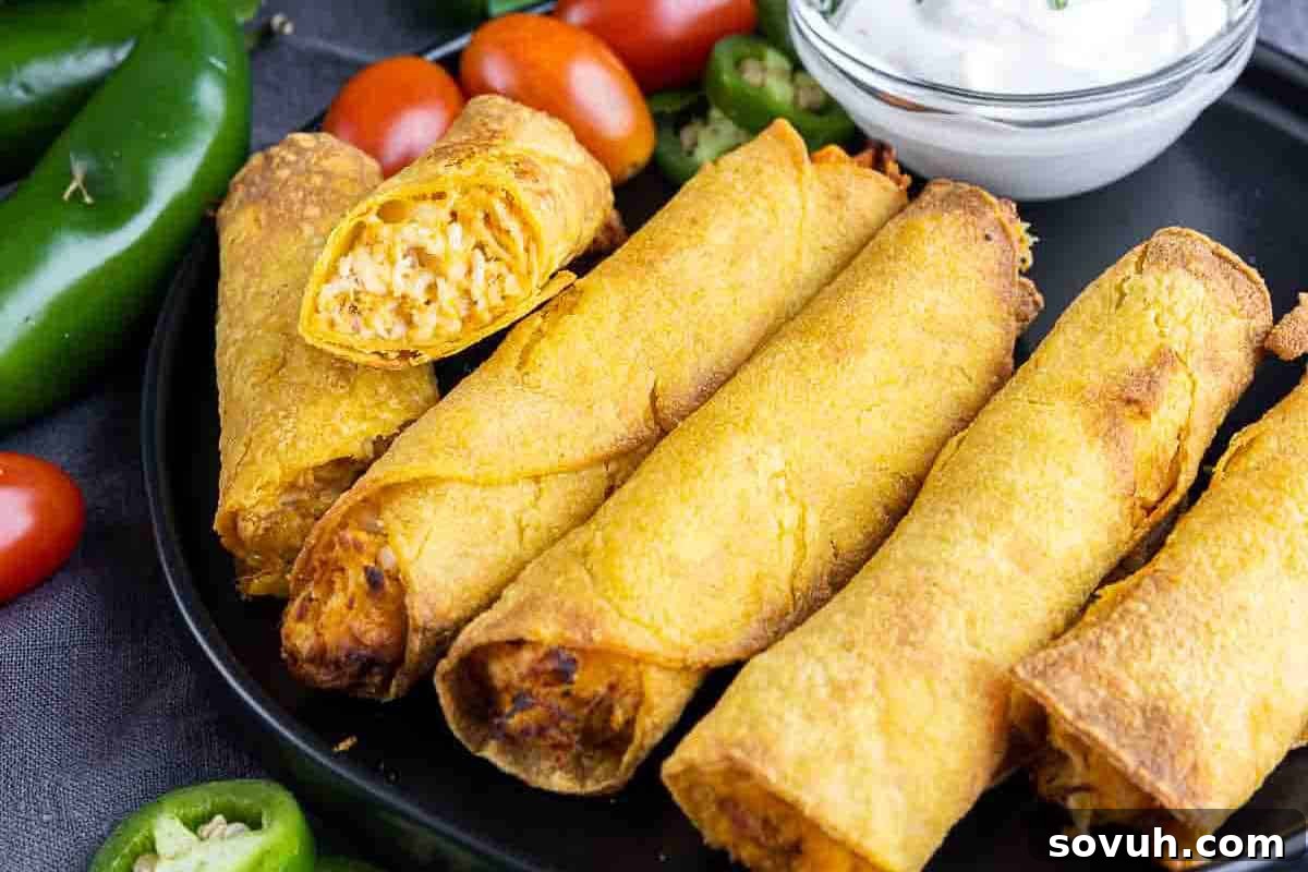 Five crispy taquitos on a black plate, one cut open to show filling. Sliced jalapeños, cherry tomatoes, and a small bowl of white dipping sauce are on the side.