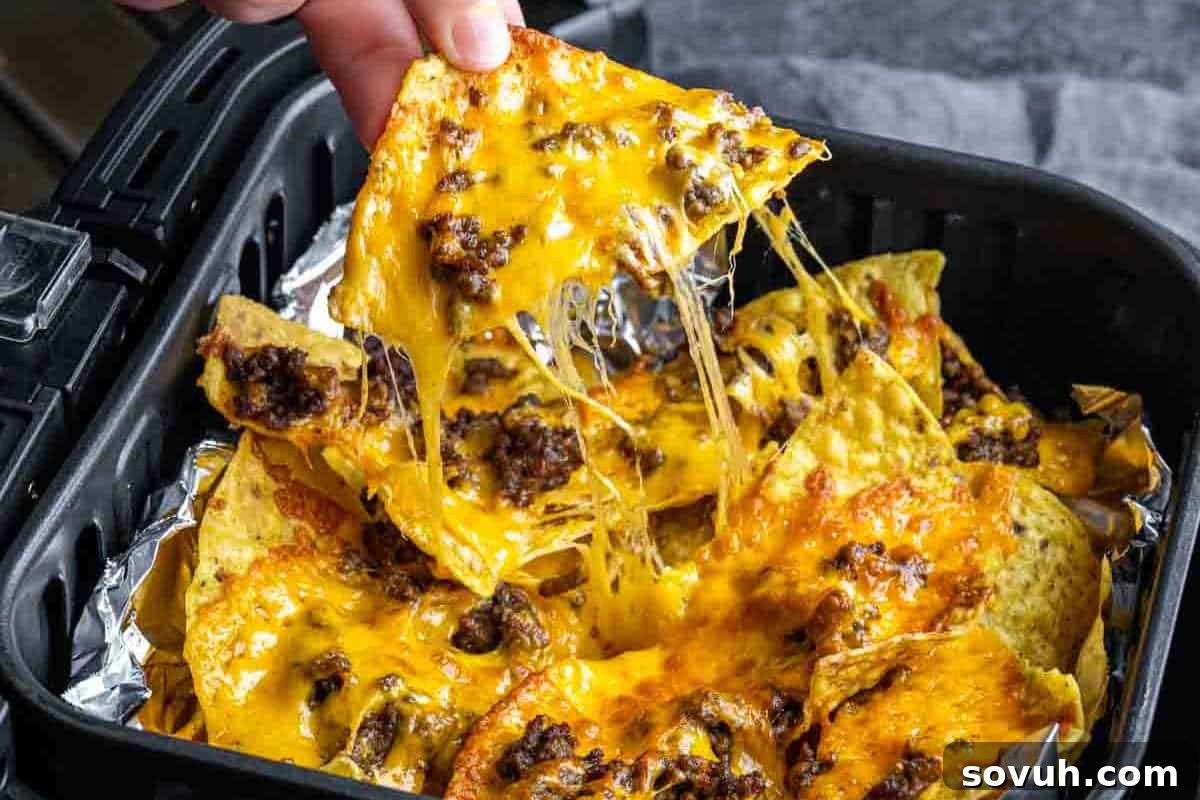A hand lifting a cheesy nacho chip topped with melted cheese and ground beef from a tray.
