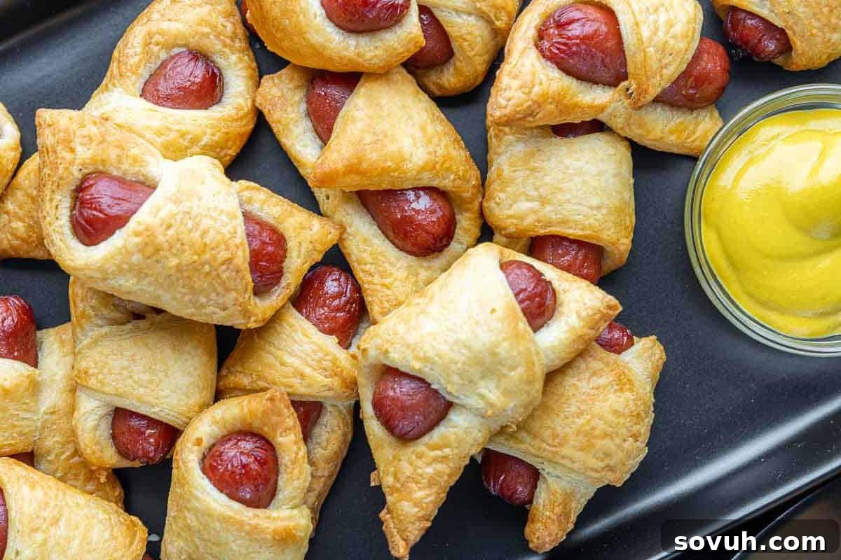 Pigs in a blanket arranged on a black platter next to a small bowl of mustard.