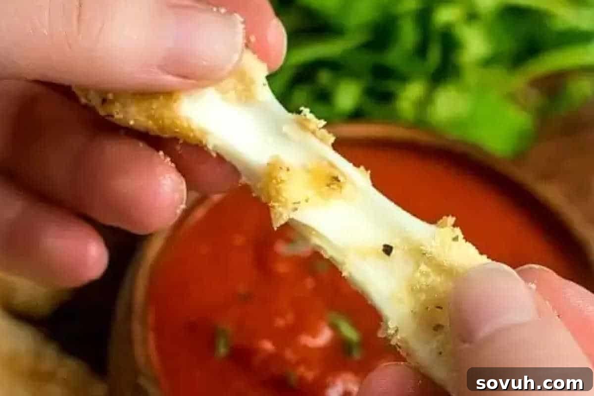 Hands pulling apart a mozzarella stick with melted cheese, above a bowl of marinara sauce and greens in the background.