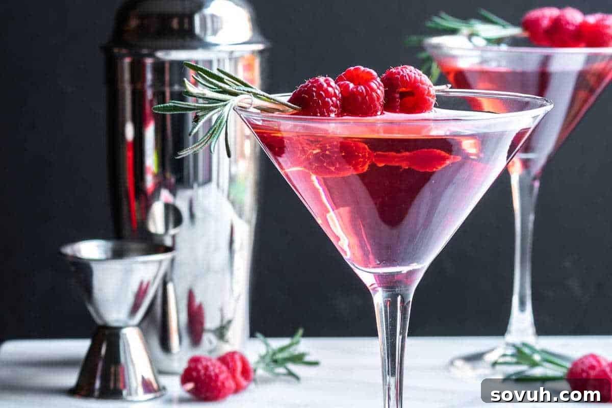Two pink cocktails with raspberries and rosemary garnish in martini glasses; a cocktail shaker and jigger in the background, setting a festive mood for a Galentine's celebration.