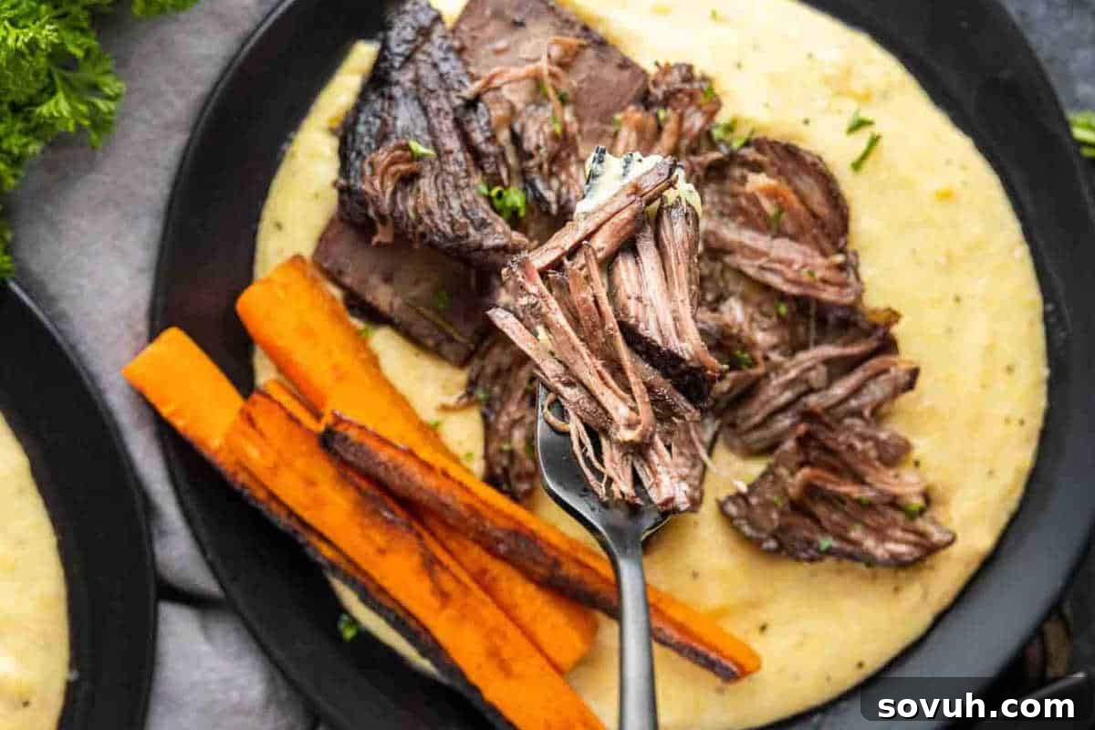 Plate of braised beef with creamy polenta and roasted carrot sticks, garnished with herbs, showing a comforting and elegant meal.
