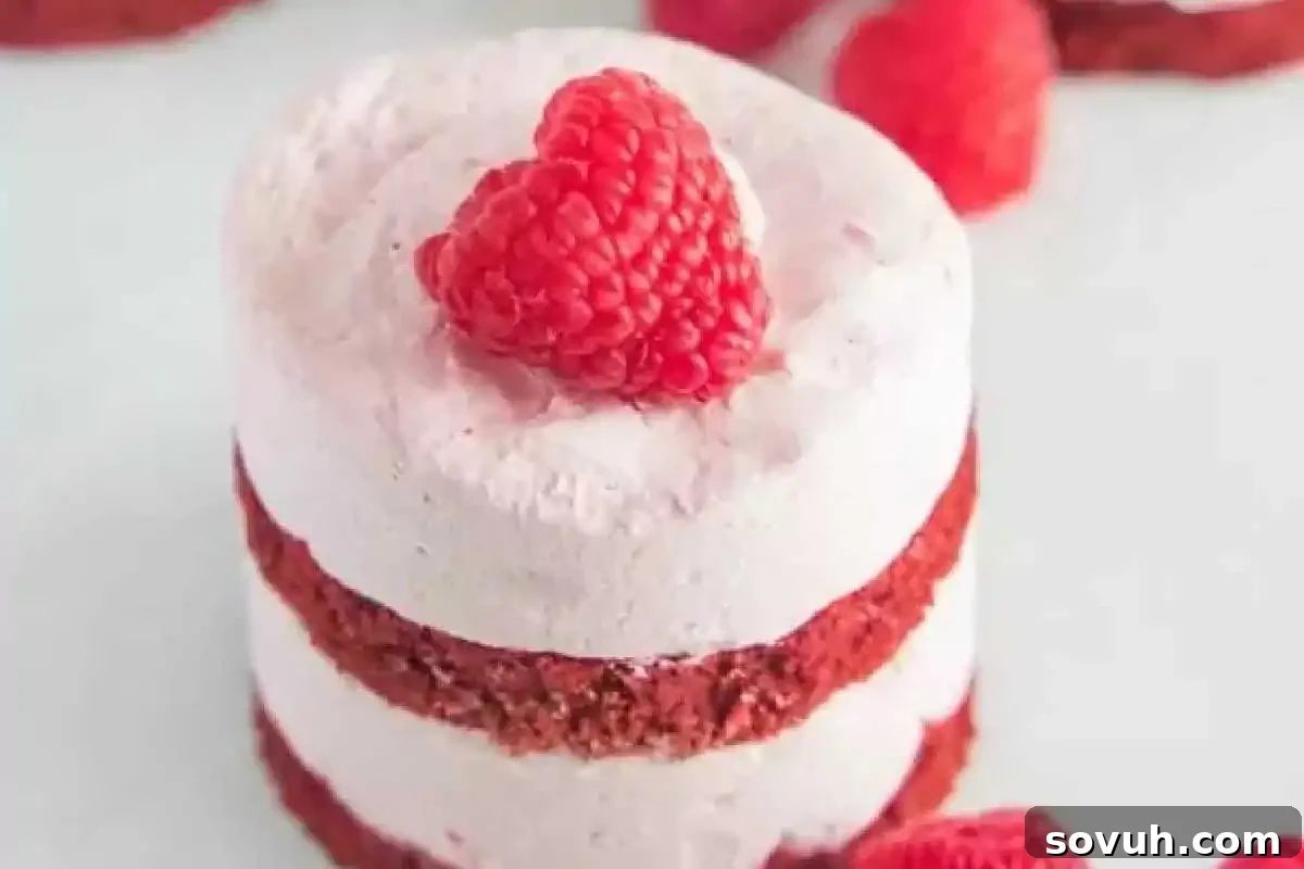 Small layered red velvet cake with white frosting, topped with a raspberry, on a white surface, perfect for a romantic or celebratory dessert.