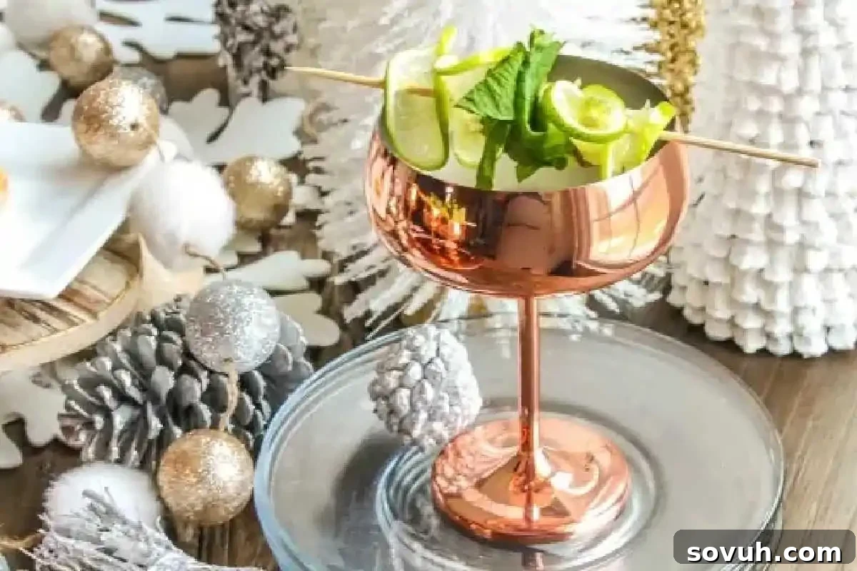 A copper cocktail glass filled with lime and mint, placed on a glass plate. Surrounded by pine cones and festive silver and white decorations, suggesting a celebratory occasion.