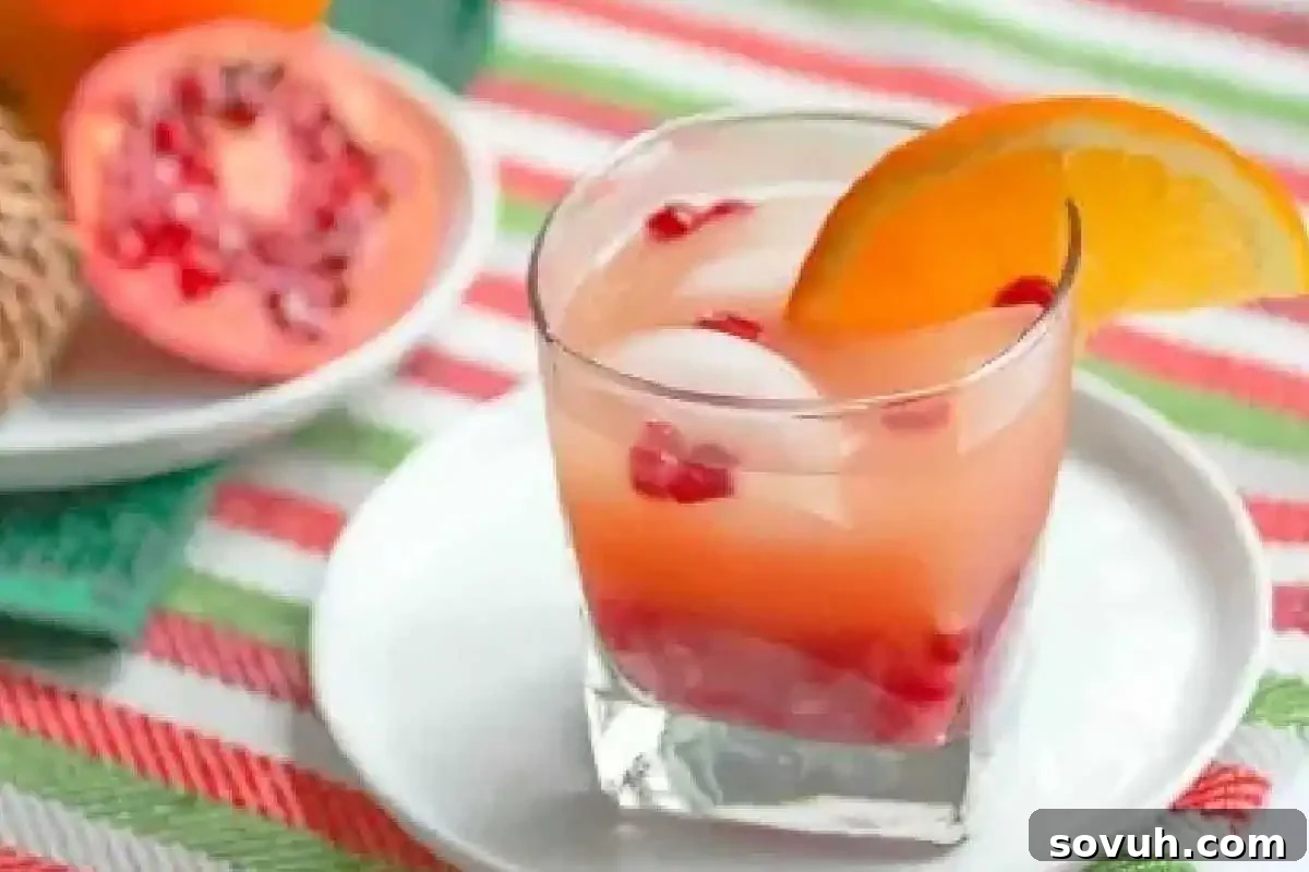 A glass of pink juice with ice cubes, pomegranate seeds, and an orange slice garnish, set on a white plate on a striped tablecloth. A halved pomegranate is in the background, highlighting fresh ingredients.