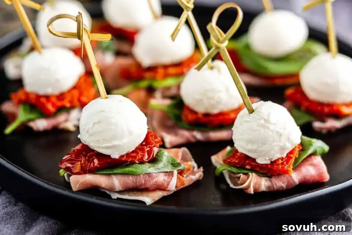 Skewered appetizers with mozzarella balls, basil, sun-dried tomatoes, and prosciutto on a black tray, perfect for a party.