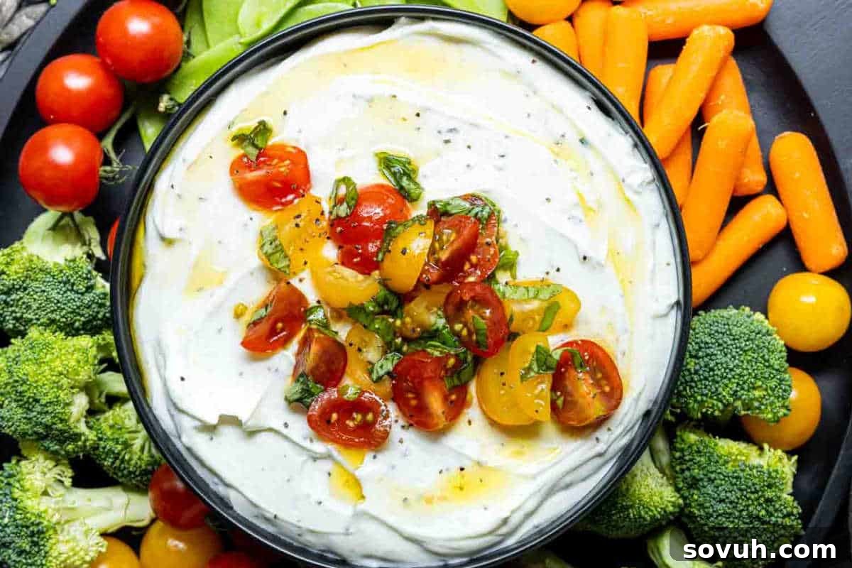 A bowl of creamy dip topped with chopped tomatoes and herbs, surrounded by broccoli, cherry tomatoes, snap peas, and baby carrots, indicating fresh ingredients.