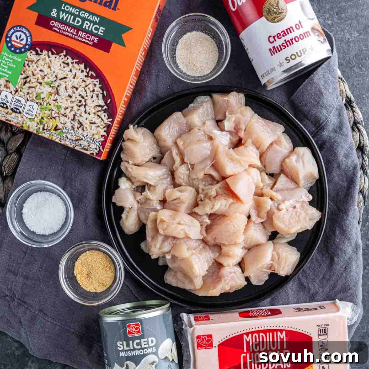A black plate with diced raw chicken surrounded by rice, a can of sliced mushrooms, cream of mushroom soup, cheddar cheese, and small bowls of spices on a textured surface.