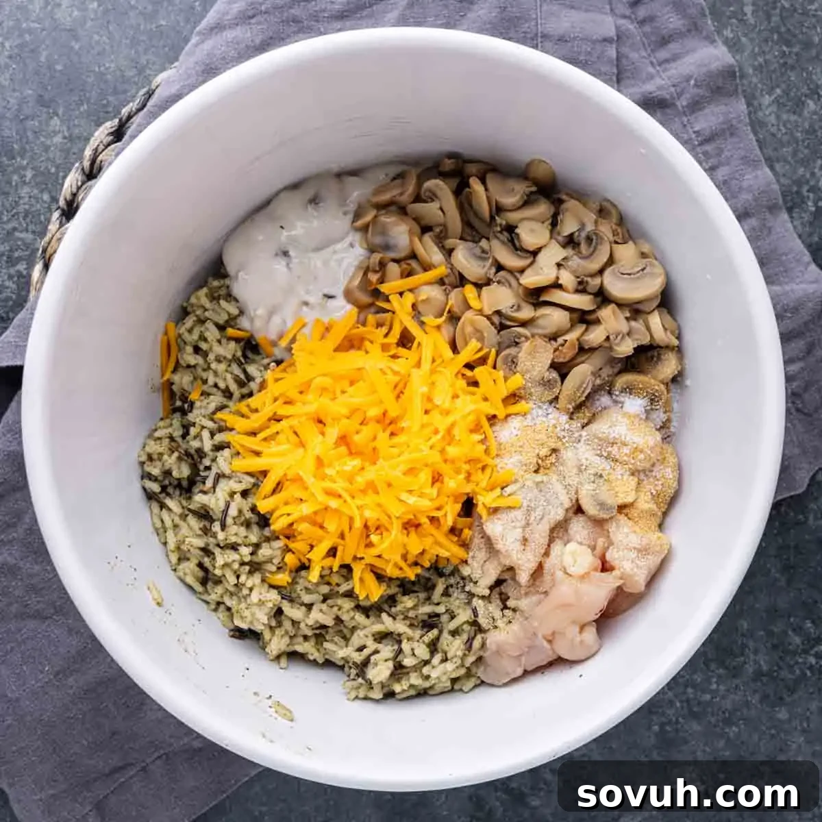 A bowl with chopped mushrooms, shredded cheese, diced chicken, sour cream, seasoned rice, and sauce on a gray cloth.