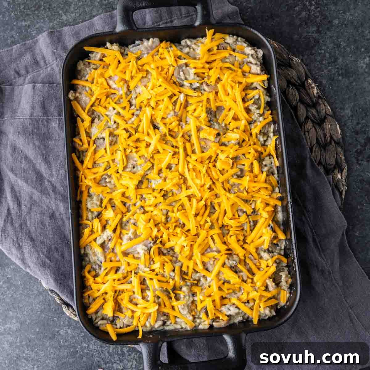 Casserole dish filled with a creamy mixture, topped with shredded cheddar cheese, placed on a dark, textured surface.