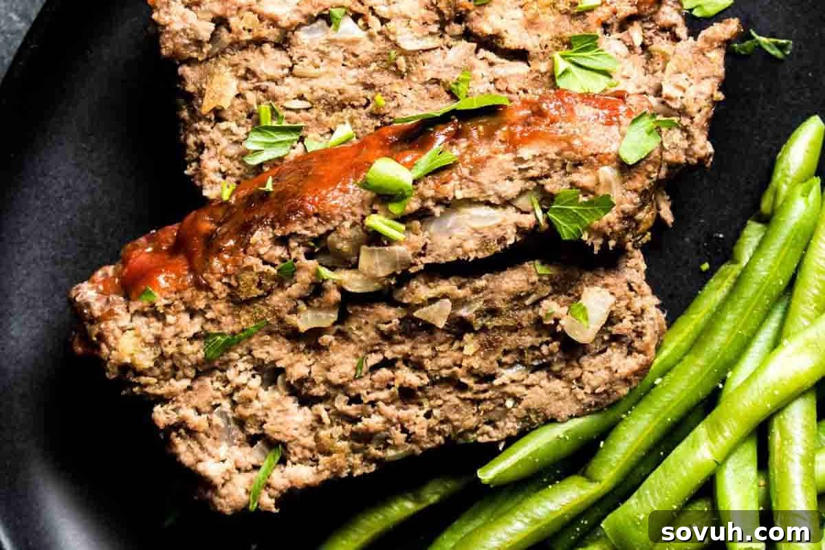 Two thick slices of delicious homemade meatloaf, glistening with a savory glaze and garnished with fresh green herbs, served next to perfectly cooked green beans on a dark, elegant plate.