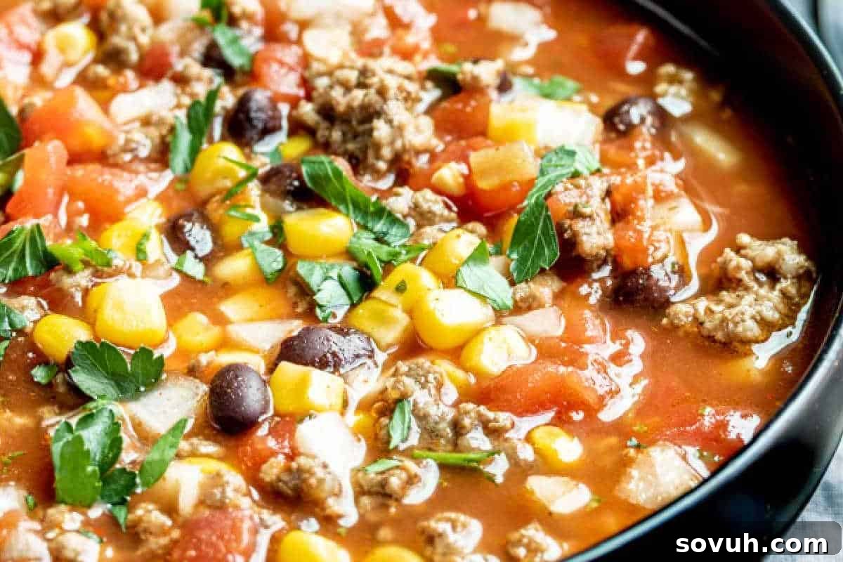 A close-up of a rustic bowl of vibrant taco soup, brimming with seasoned ground beef, golden corn kernels, black beans, diced tomatoes, and garnished with fresh green parsley, ready to be enjoyed.
