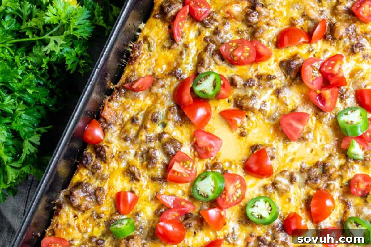 A vibrant Taco Casserole in a baking dish, generously topped with melted cheese, seasoned ground meat, fresh chopped tomatoes, and sliced jalapeños, with fresh parsley artfully arranged on the side.
