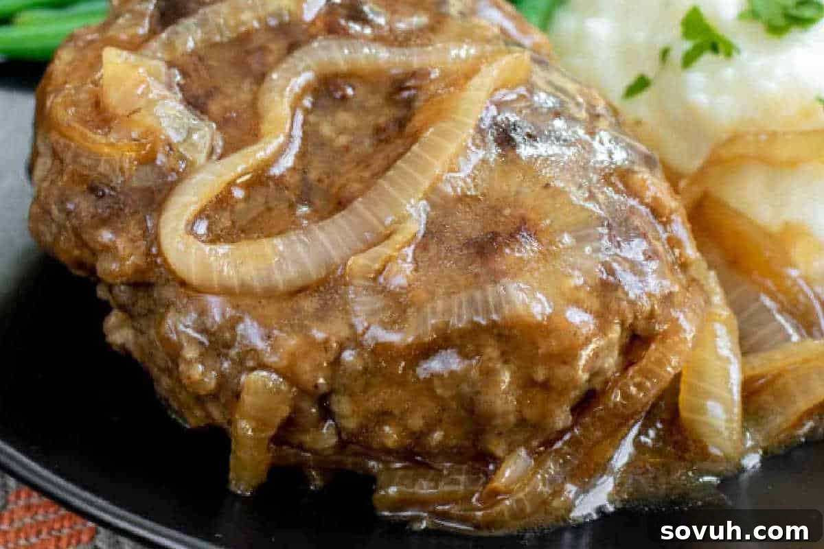 A classic Salisbury steak patty topped with caramelized onions and rich brown gravy, served alongside a scoop of creamy mashed potatoes on a sleek black plate.