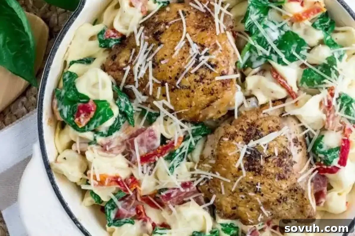 A white pan filled with a vibrant One Pot Creamy Chicken Tortellini dish, featuring perfectly cooked tortellini, fresh spinach, sweet sun-dried tomatoes, and two pieces of beautifully seasoned grilled chicken, all generously topped with grated Parmesan cheese.