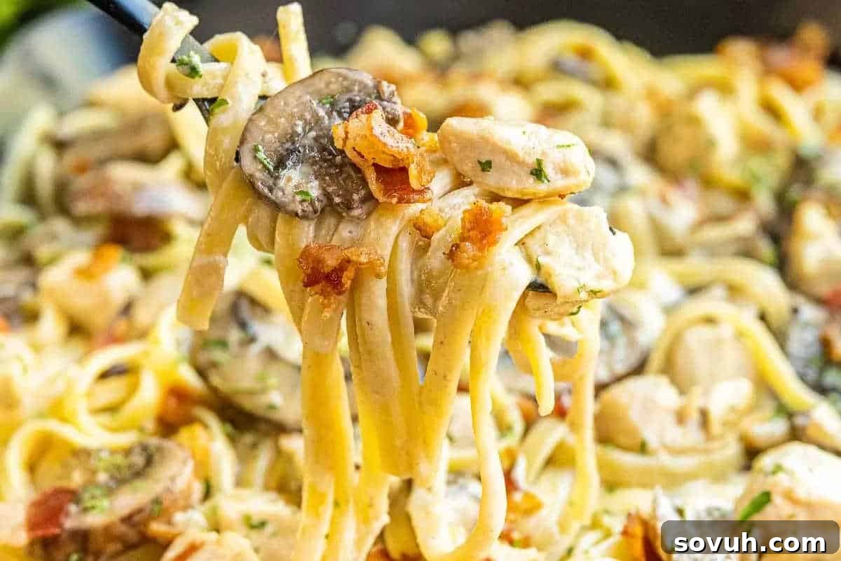 A close-up of a fork lifting a generous portion of creamy pasta, showcasing tender mushrooms, succulent chicken pieces, and crispy bacon bits, highlighting the rich textures and savory ingredients.
