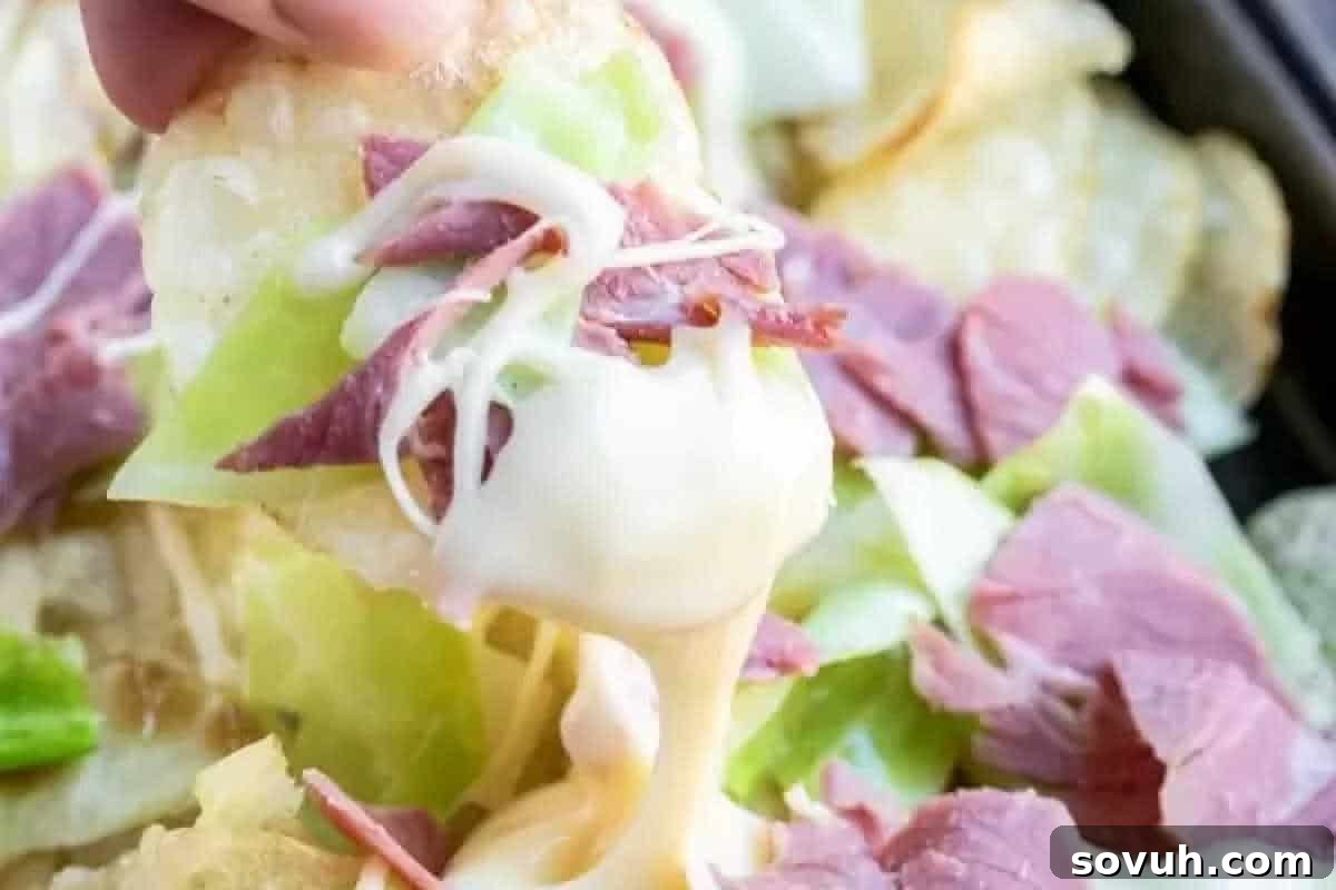 A hand skillfully lifts a single chip loaded with melted cheese, tender sliced corned beef, and vibrant cabbage, poised above a generous plate of equally delicious Irish Nachos.