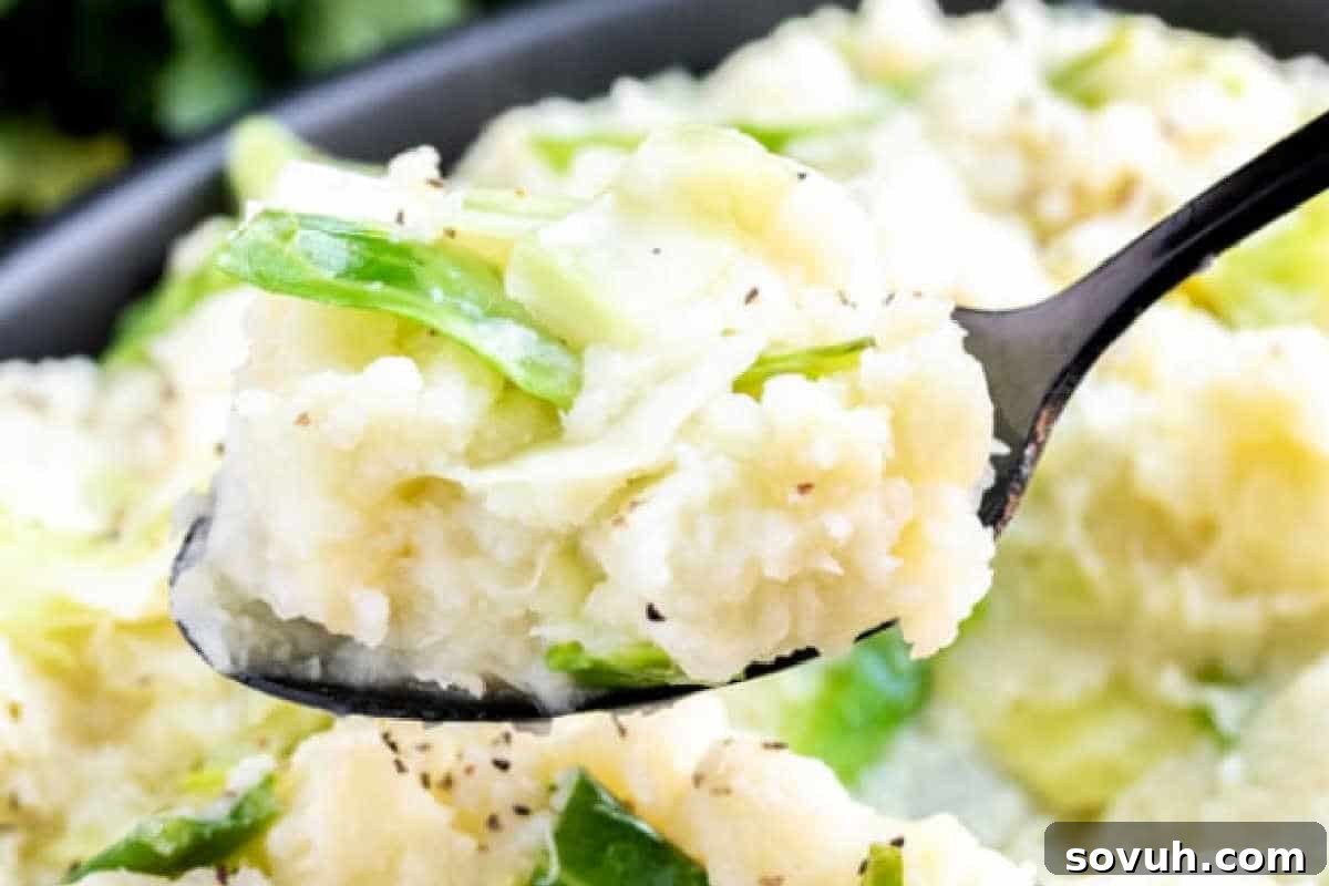 A serving spoon lifting a portion of creamy mashed potatoes generously mixed with vibrant green cabbage and a sprinkle of freshly ground black pepper.