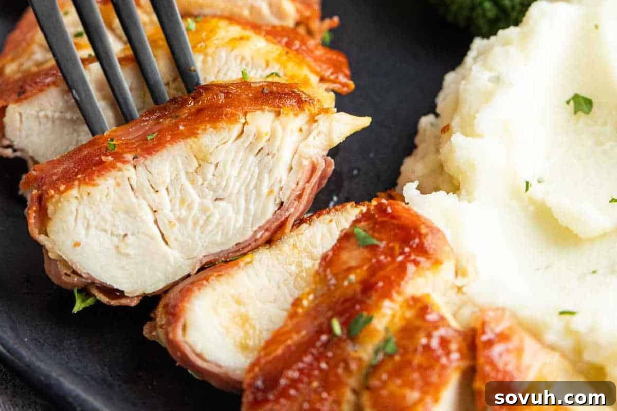 Sliced chicken wrapped in bacon on a plate, next to a serving of mashed potatoes. A fork holds a piece of chicken.