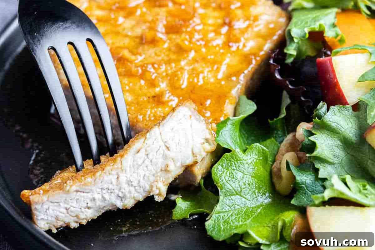 A fork slices into a piece of glazed pork chop on a plate, accompanied by a fresh salad with leafy greens and apple slices.