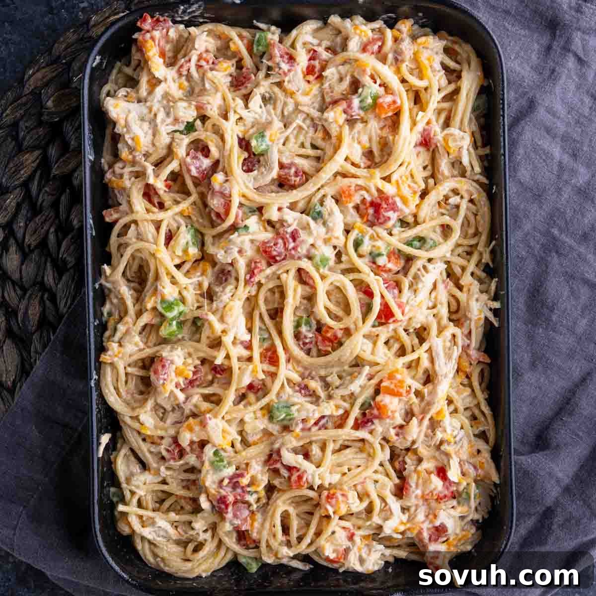 Rectangular baking dish filled with creamy pasta, mixed with visible chunks of red and green bell peppers and tomatoes, resting on a dark cloth.