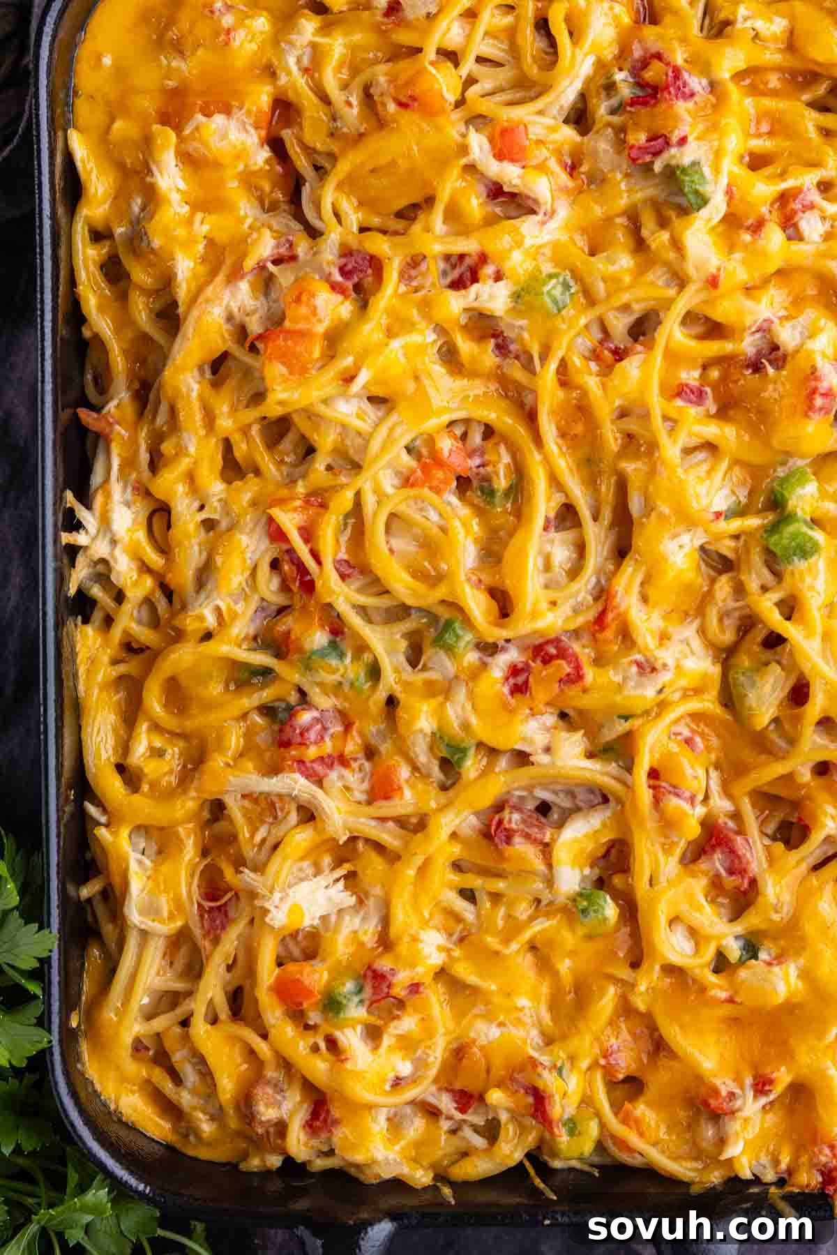 Close-up of a baked spaghetti casserole topped with melted cheese, diced vegetables, and bacon bits, in a black rectangular dish.